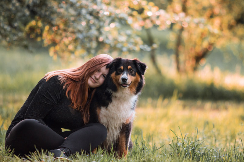 Besitzerin eines black tri Australian Shepherds sitzt mit ihrem Hund auf einer Wiese und lehnt sich an ihn an