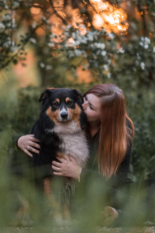 Besitzerin eines black tri Australian Shepherds sitzt neben ihrem Hund, umarmt ihn und gibt ihm ein Küsschen